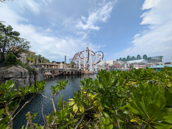 Theme park scene featuring a roller coaster with lush greenery and a water body.