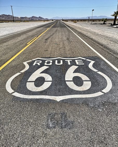 A weathered Route 66 sign painted on a deserted road with mountains in the background.