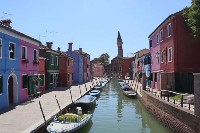 Colourful houses lining a canal in Burano, Italy, with a bell tower in the background.