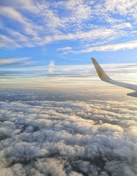 Vueling airplane wing above fluffy clouds with a bright blue sky and wispy clouds.