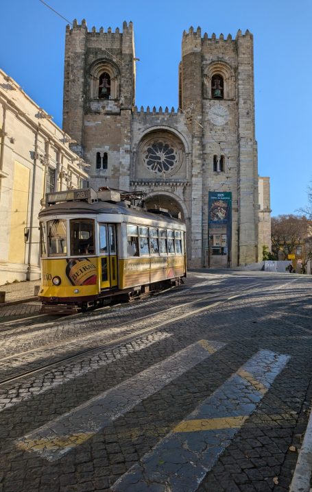 Vintage yellow tram passing by a historic building with towers and a large clock.