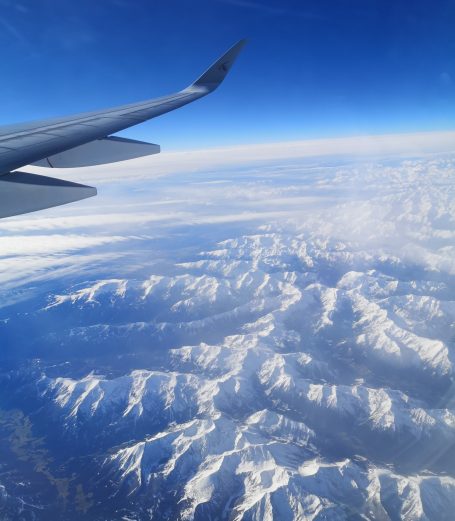 Snow-capped mountains below, seen from an airplane wing against a blue sky.