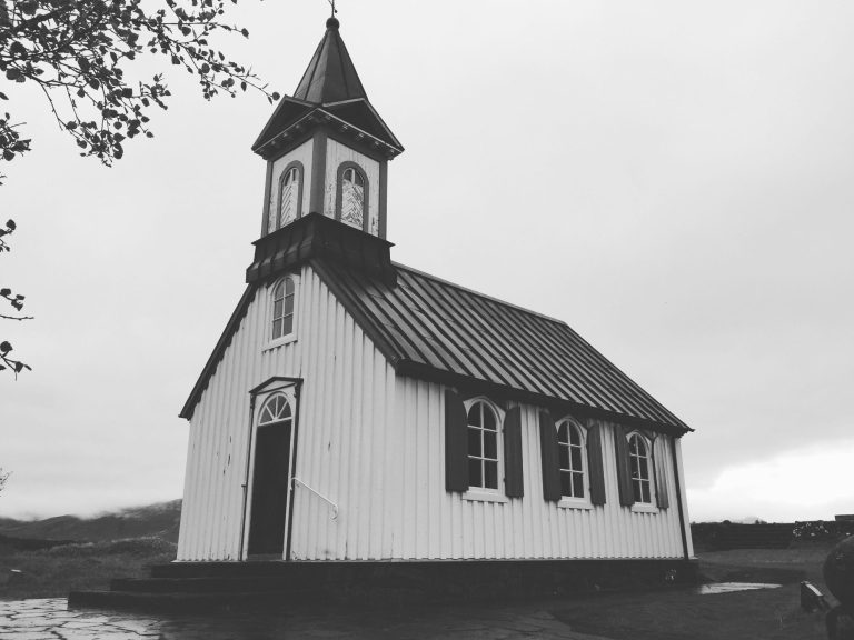 Small white church with a steep roof and tall spire set against a grey sky.