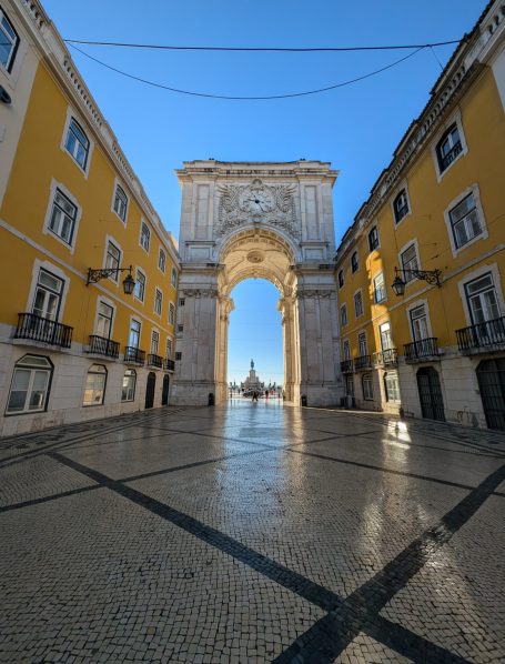 Archway leading to a square, framed by yellow buildings and a clear blue sky.